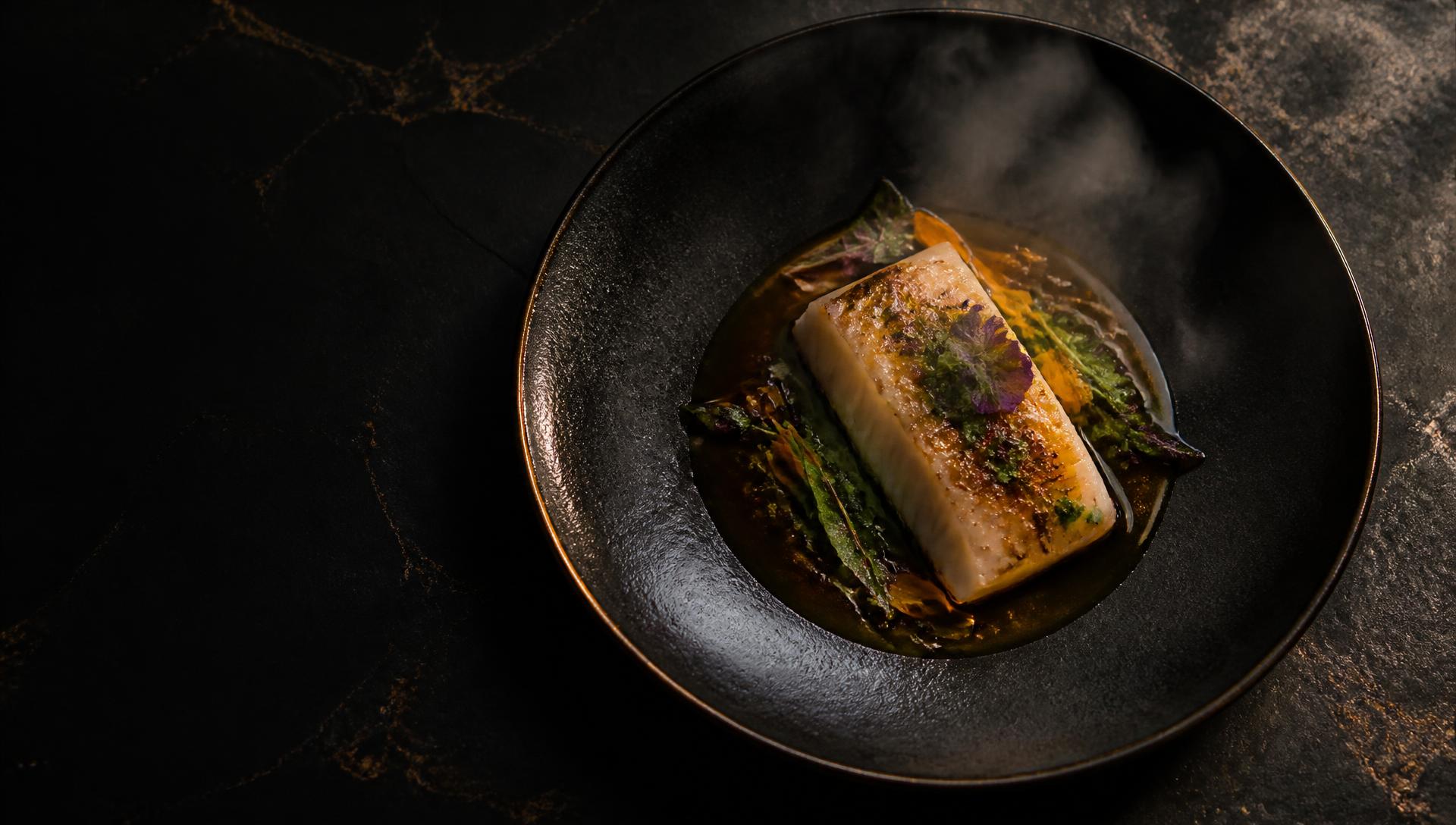 Plato de cocina de autor: pescado blanco con salsa oscura, hierbas y flores comestibles sobre mesa de piedra